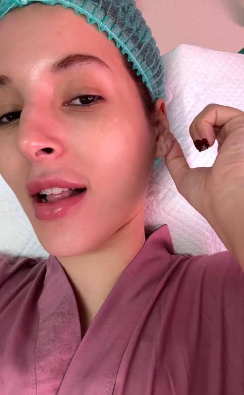 Seorang perempuan mengenakan pakaian medis berwarna merah muda dan penutup kepala hijau muda sambil memegang telinganya di ruang perawatan.