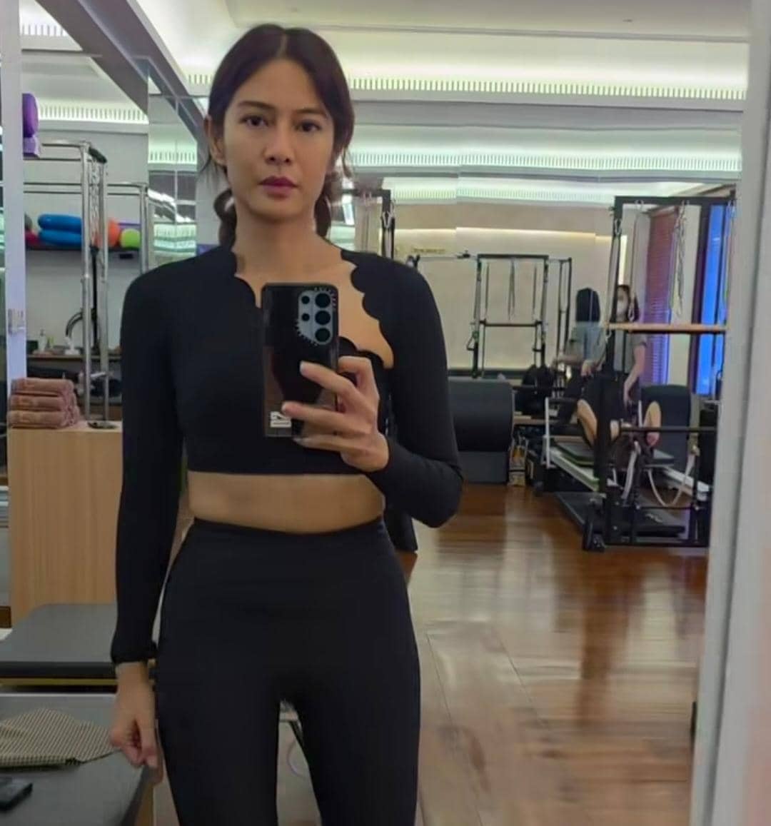 Seorang perempuan mengenakan pakaian olahraga hitam berpose di depan cermin di studio pilates dengan peralatan latihan di belakangnya.