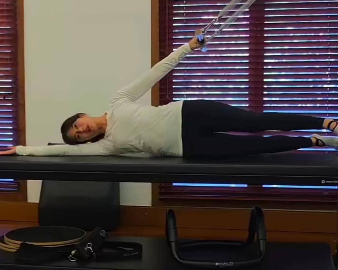 Seorang perempuan mengenakan pakaian olahraga putih dan hitam sedang melakukan latihan pilates di atas reformer di dalam ruangan.