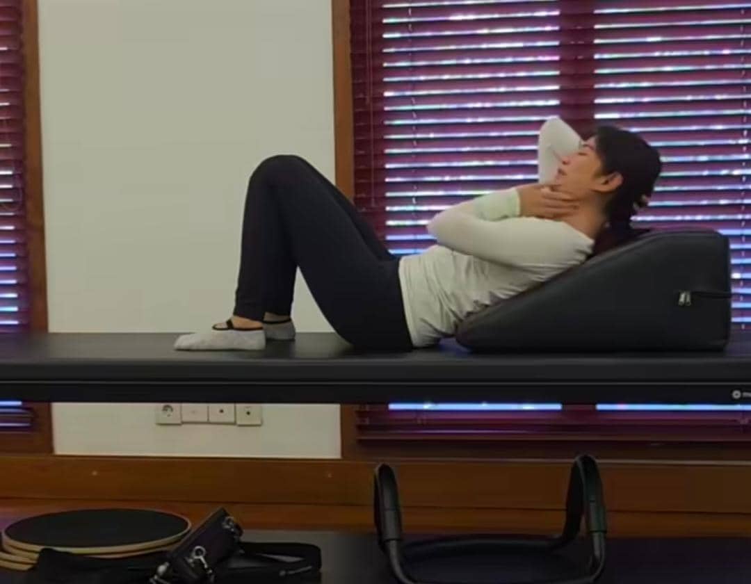 Seorang wanita mengenakan pakaian olahraga putih dan hitam sedang melakukan gerakan pilates di atas matras di dalam ruangan dengan jendela kayu.