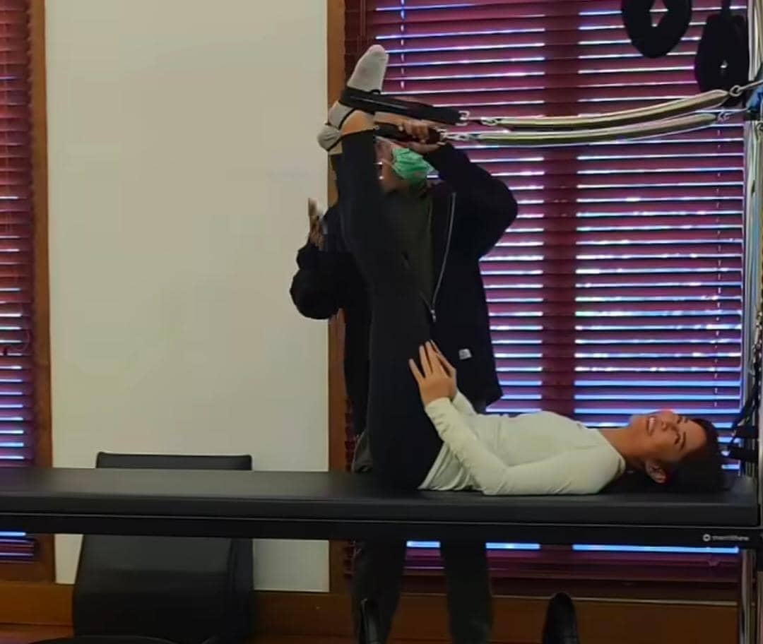 Seorang wanita berbaring di alat pilates sambil dibantu instruktur yang mengenakan masker di dalam ruangan dengan jendela kayu.