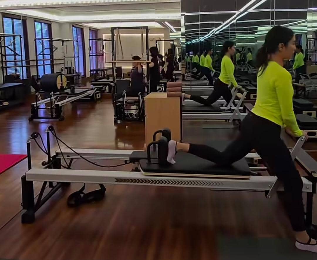 Seorang wanita mengenakan pakaian olahraga berwarna hijau neon sedang melakukan latihan pilates di studio dengan peralatan lengkap dan cermin besar.