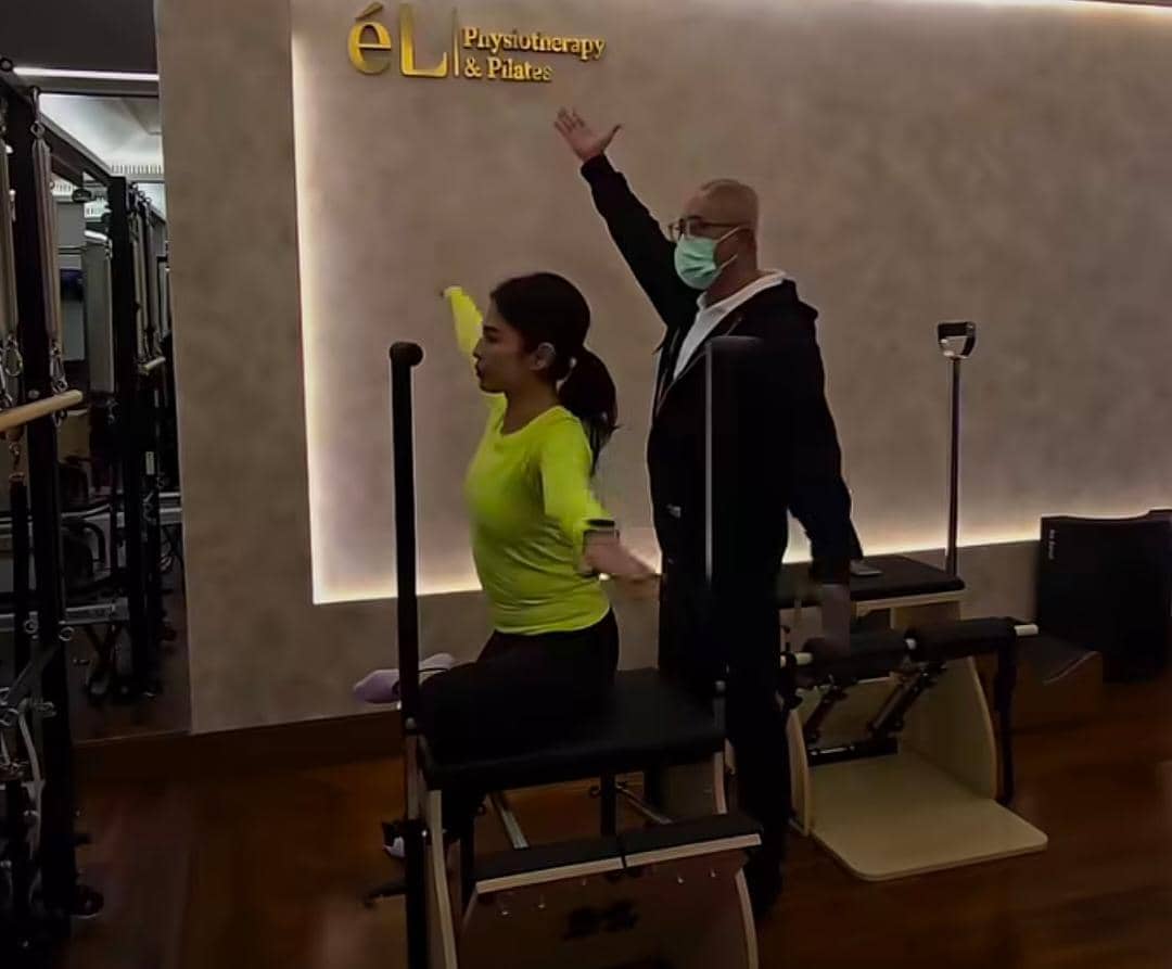 Seorang wanita berolahraga pilates dengan instruktur di studio bertuliskan ÉL Physiotherapy & Pilates, keduanya mengangkat tangan.