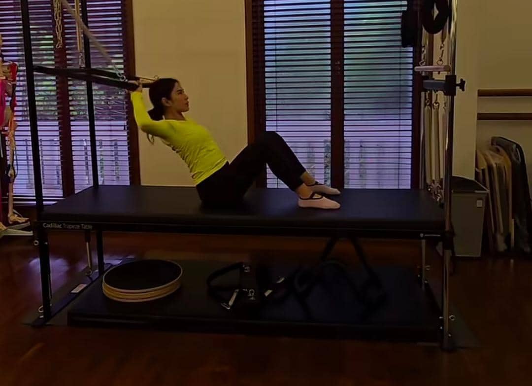 Seorang wanita mengenakan atasan kuning dan celana hitam sedang melakukan latihan pilates di atas alat reformer di ruangan berpenerangan alami.