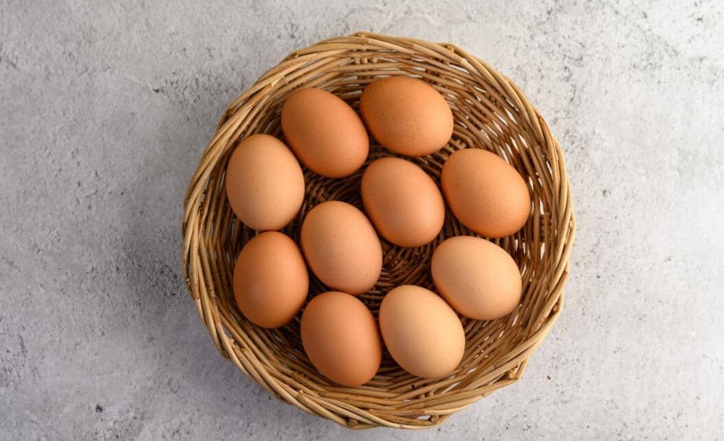 Sepuluh butir telur ayam negeri berwarna cokelat tersusun rapi di dalam keranjang rotan di atas permukaan abu-abu.
