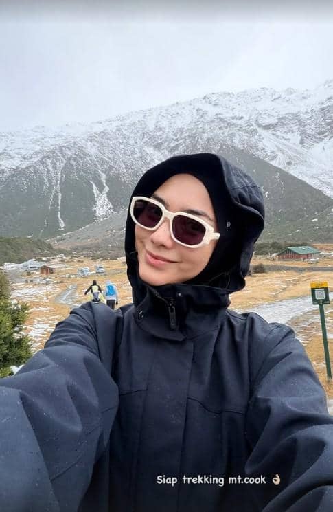 Seorang perempuan berjaket hitam dan berkacamata putih berpose selfie di area pegunungan bersalju Gunung Cook, Selandia Baru.