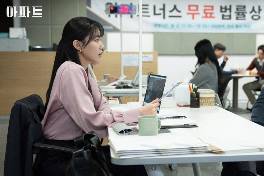 Seorang wanita duduk di meja kantor dengan laptop terbuka dan dokumen di tangannya, suasana ruang kerja modern dengan beberapa orang di latar belakang.