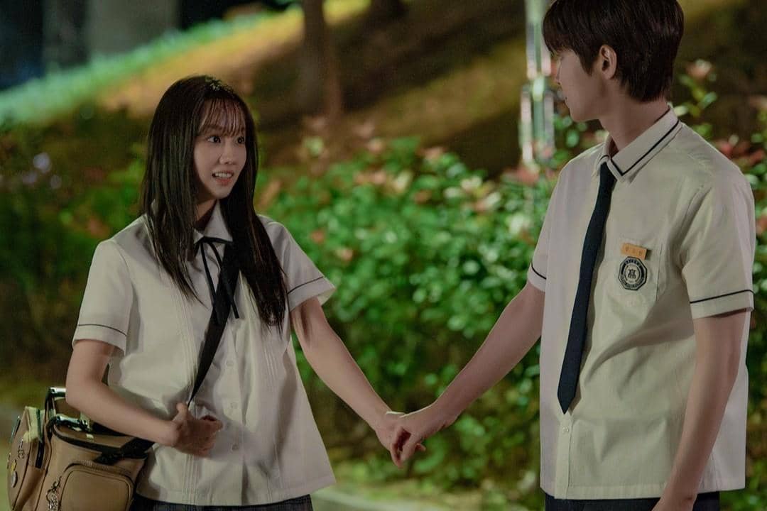 Dua siswa berseragam sekolah berdiri saling berhadapan di luar ruangan pada malam hari sambil berpegangan tangan dengan ekspresi hangat.