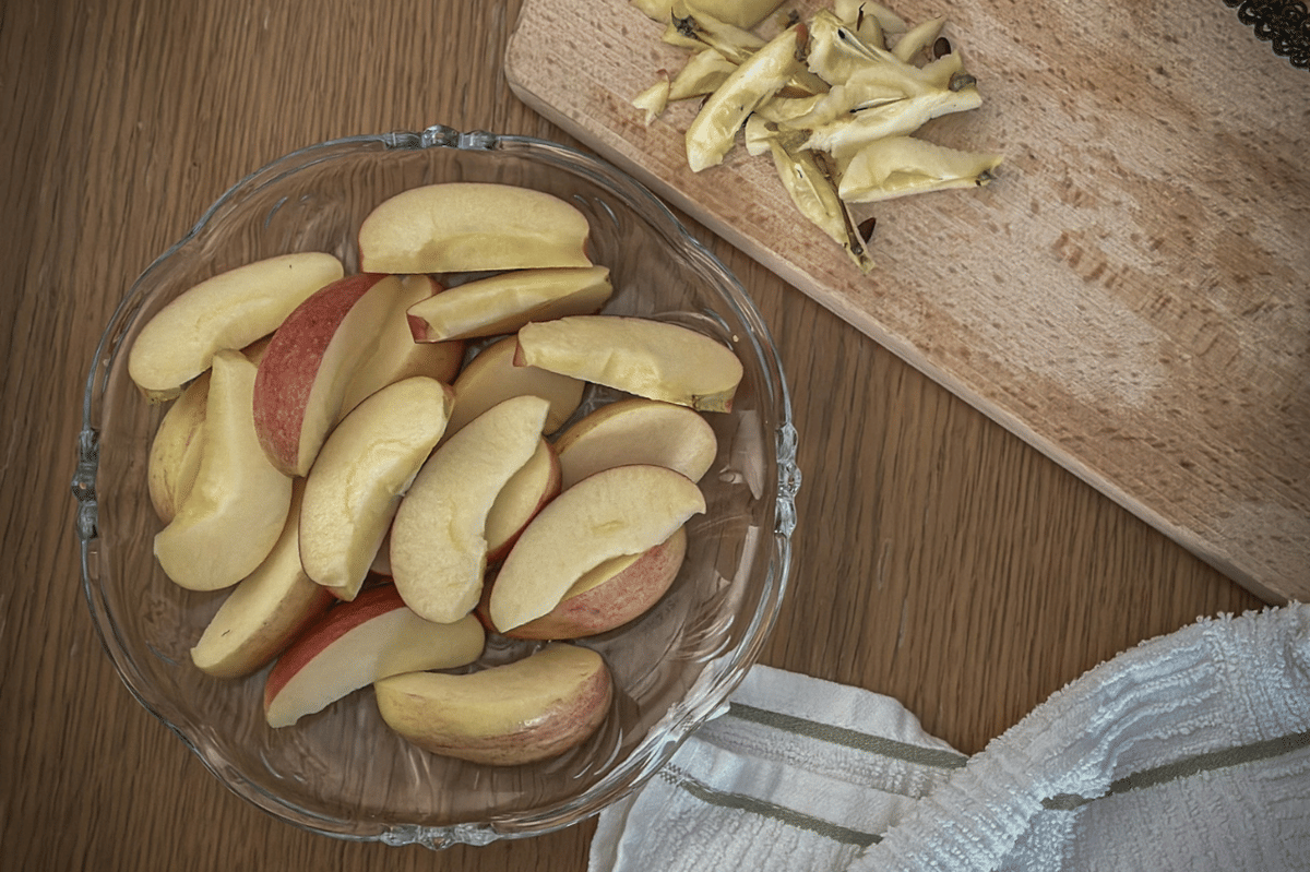 Irisan apel segar dalam mangkuk kaca di atas meja kayu dengan talenan berisi sisa potongan apel dan kain dapur putih di sampingnya.