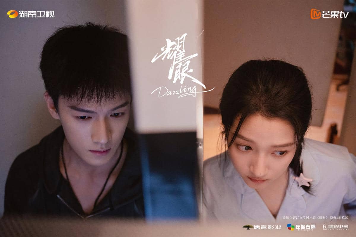 Dua pemeran utama drama China Dazzling duduk bersebelahan di depan layar komputer dengan ekspresi serius dalam suasana redup.