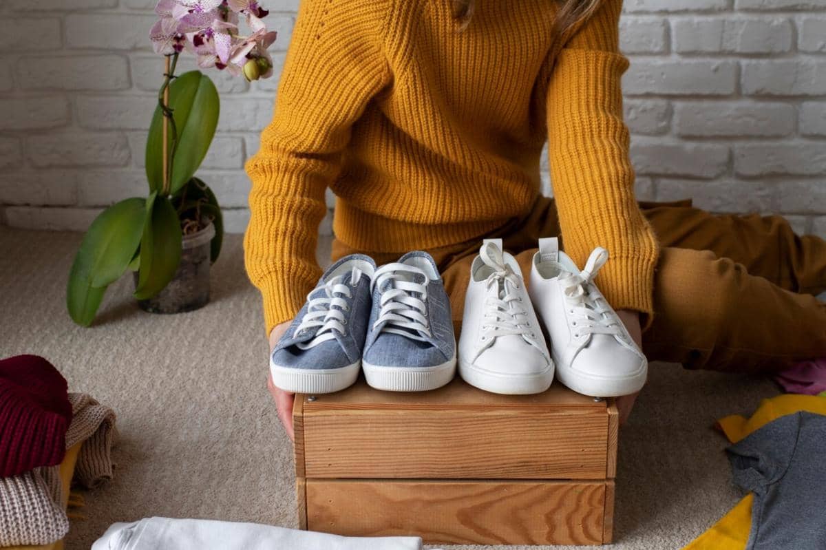 Seseorang mengenakan sweter kuning duduk di lantai sambil menata dua pasang sepatu kasual di atas kotak kayu dengan latar dinding bata putih.