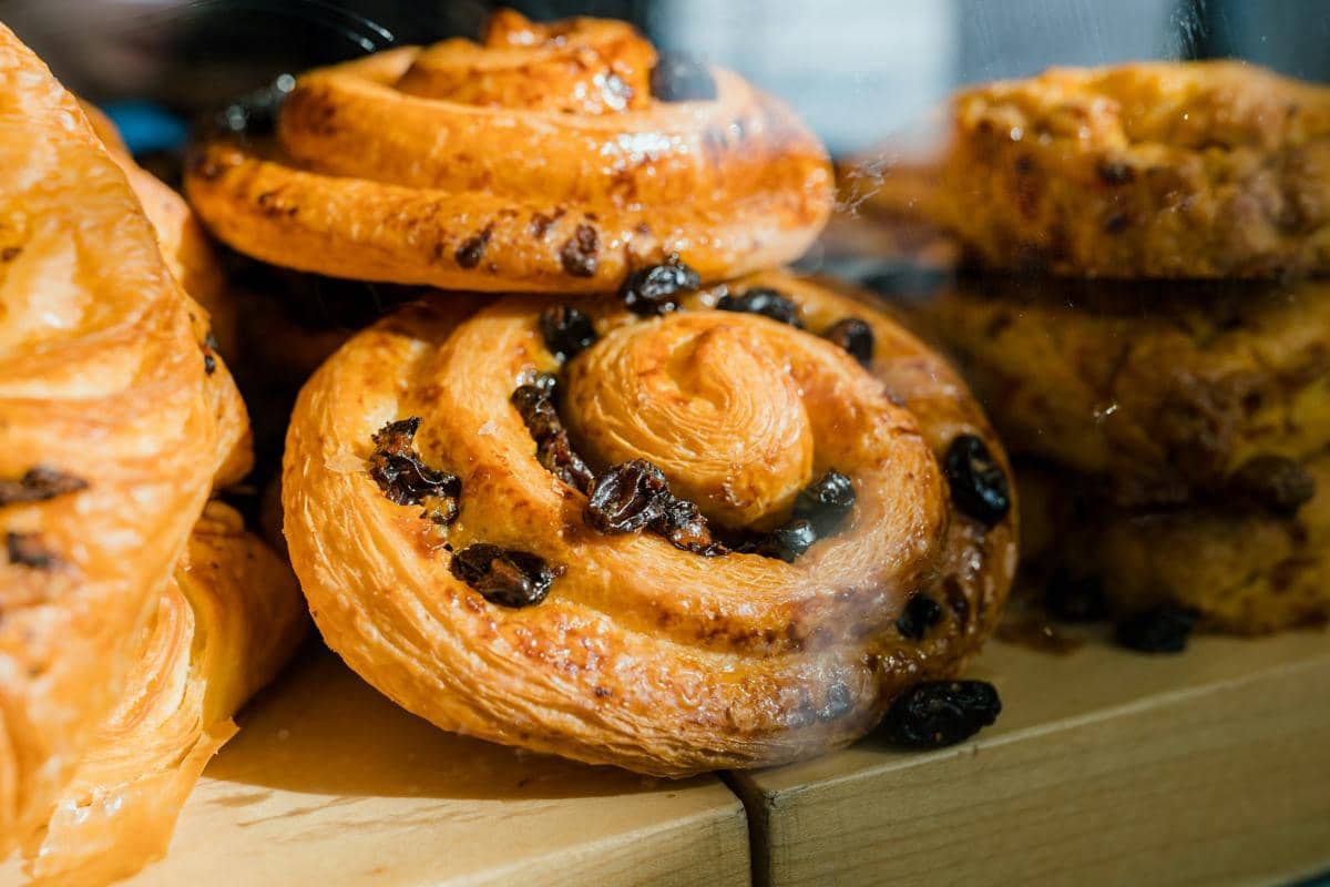 Beberapa cinnamon roll kismis berwarna keemasan dengan tekstur renyah dan kilau gula di permukaannya tersusun di atas meja kayu.
