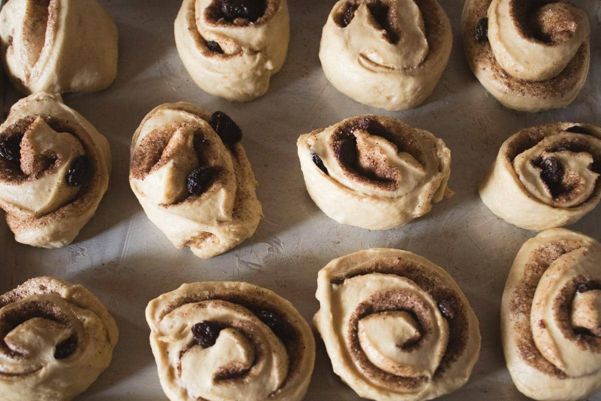 Adonan cinnamon roll kismis yang belum dipanggang tersusun rapi di atas loyang dengan taburan kayu manis dan gula.