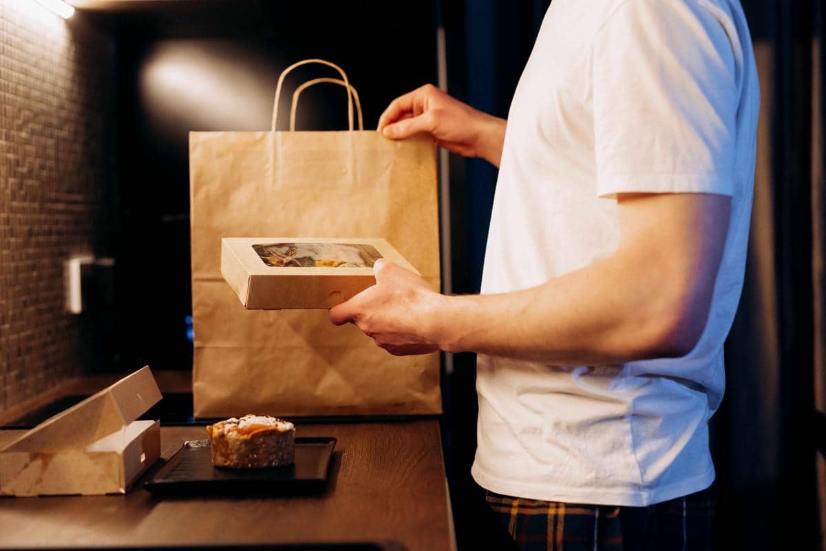 Seseorang mengenakan kaus putih memegang kotak makanan di dapur dengan tas kertas besar di meja, menggambarkan aktivitas pesan makanan online.
