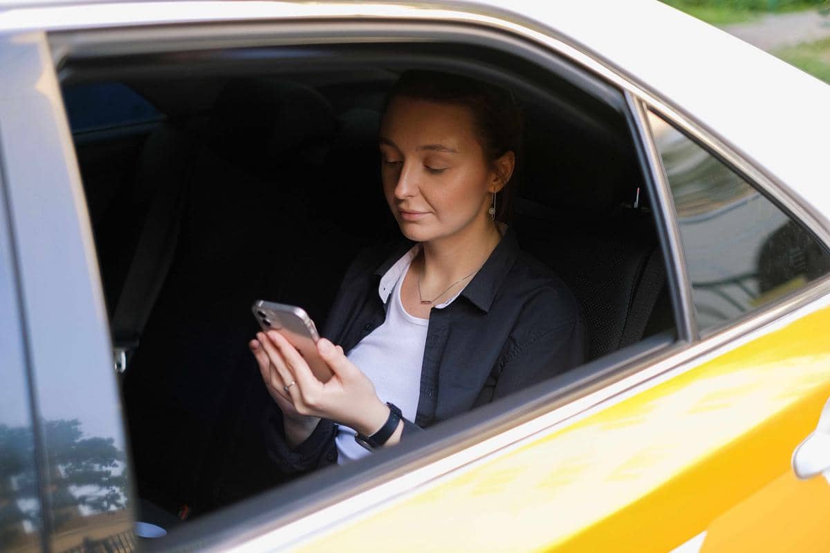 Seorang wanita duduk di kursi belakang mobil berwarna kuning sambil melihat ponsel, menggambarkan penggunaan transportasi online.