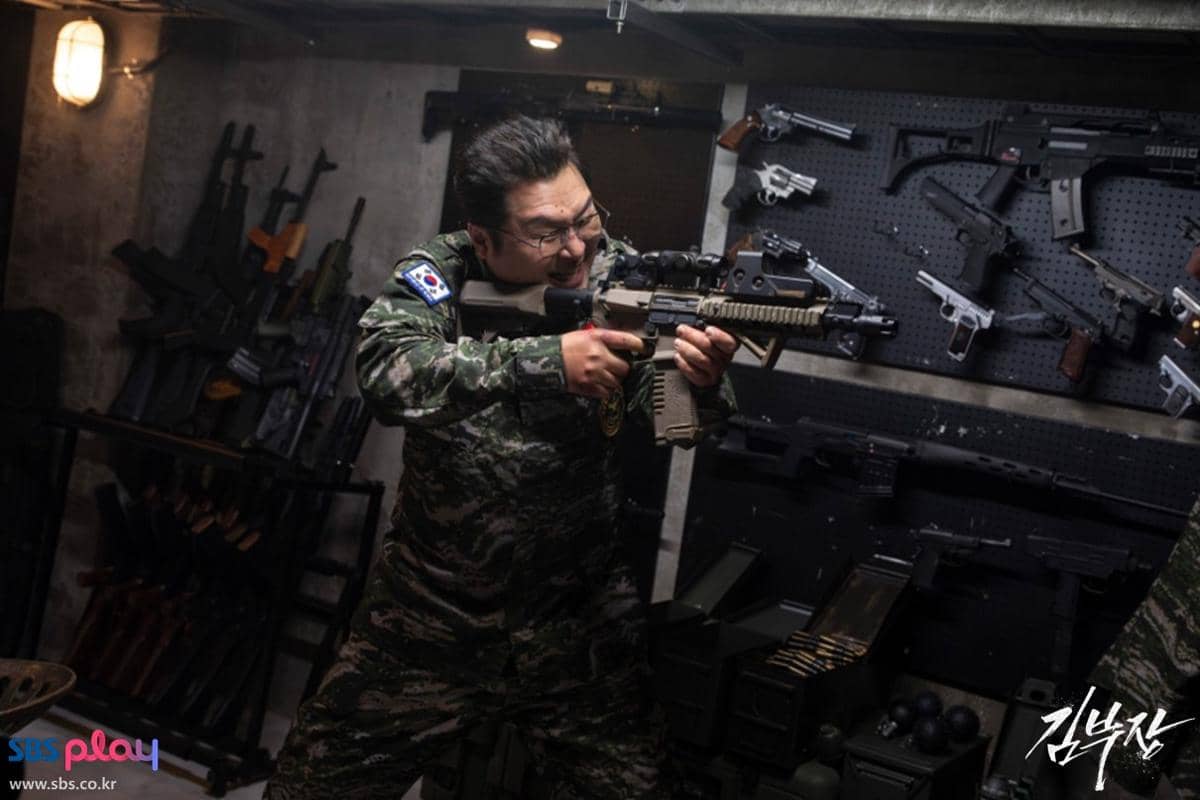 Seorang pria berseragam militer memegang senapan serbu di ruangan penuh senjata api dalam adegan drama Agent Kim Reactivated.