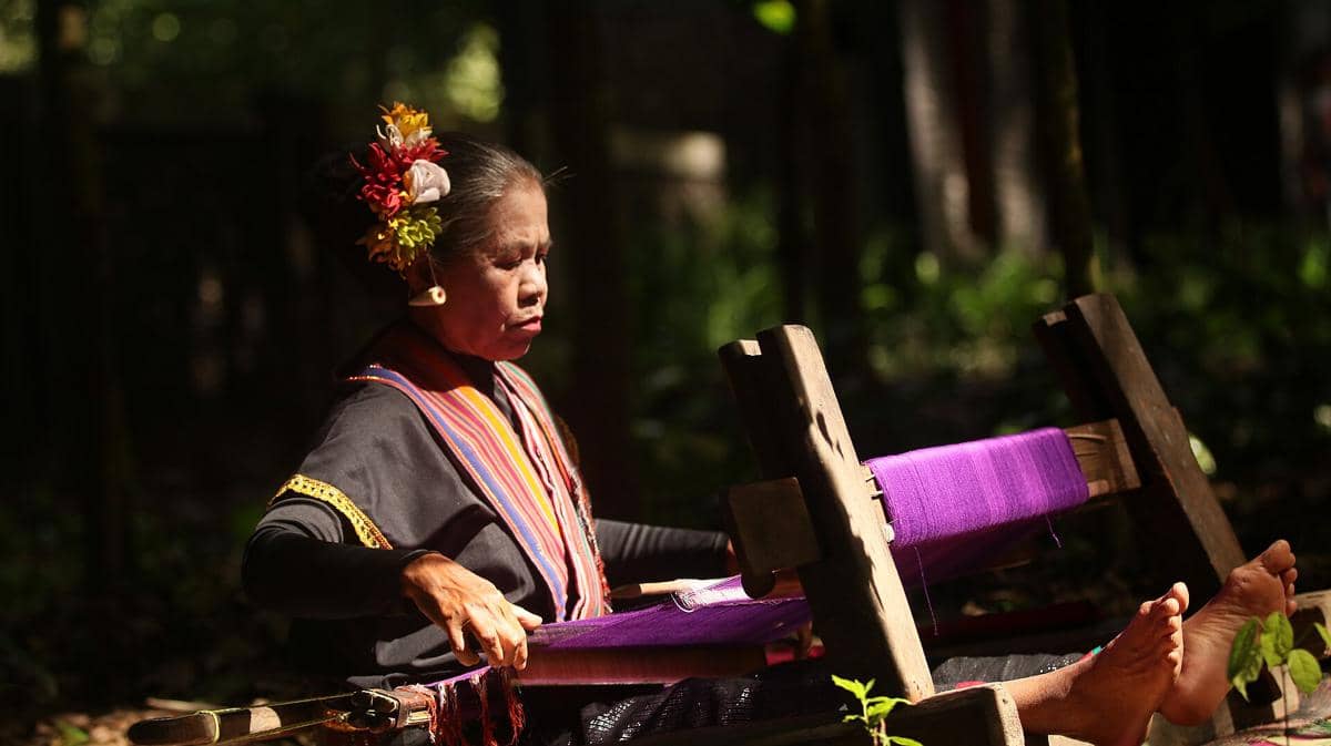 Seorang nenek Sasak tengah menenun kain tradisional dengan alat tenun dalam potret aktivitas sehari-hari.