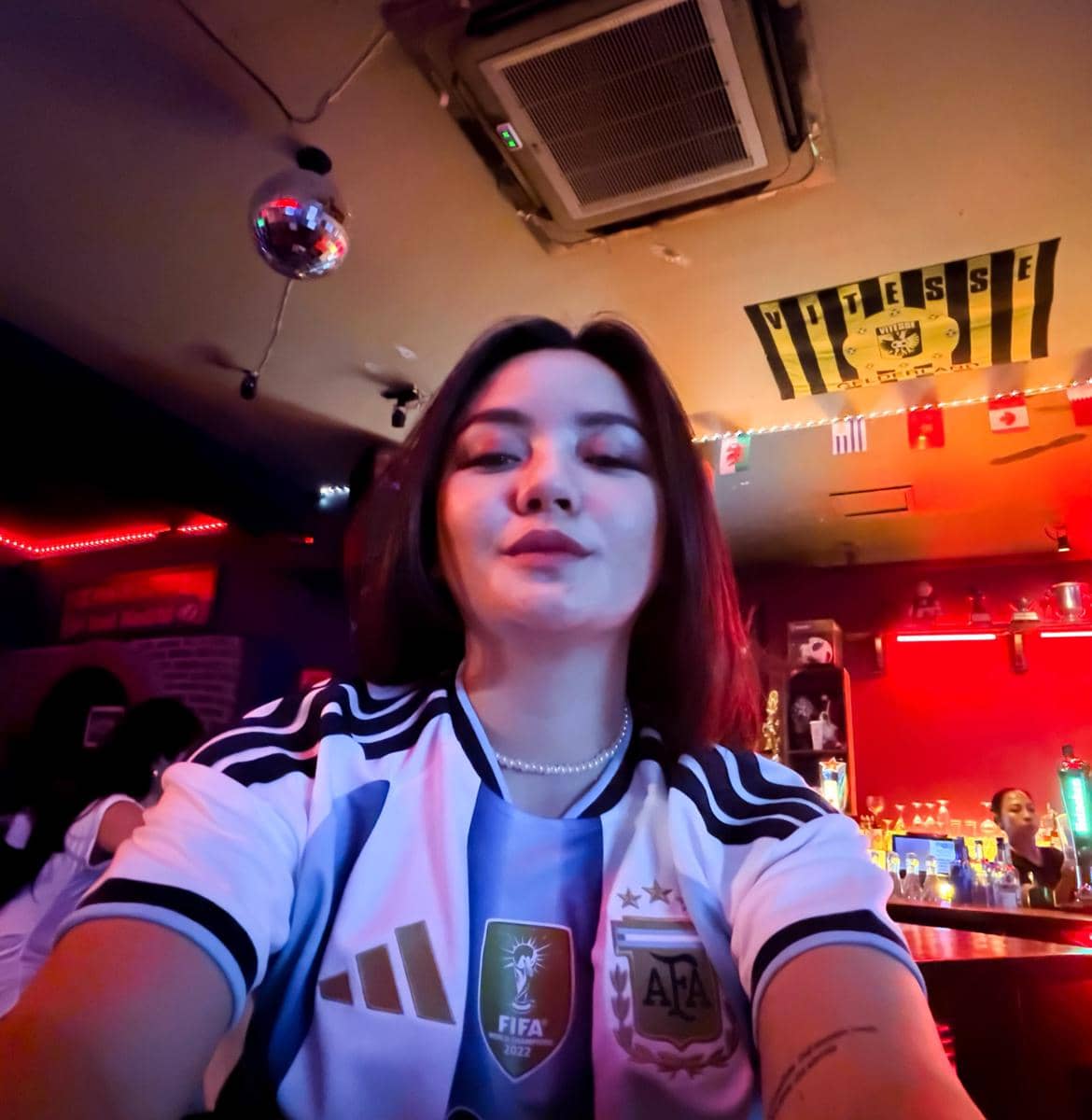 Seorang perempuan mengenakan jersey tim nasional Argentina berpose di dalam bar dengan pencahayaan neon saat acara nonton bareng Piala Dunia 2026.