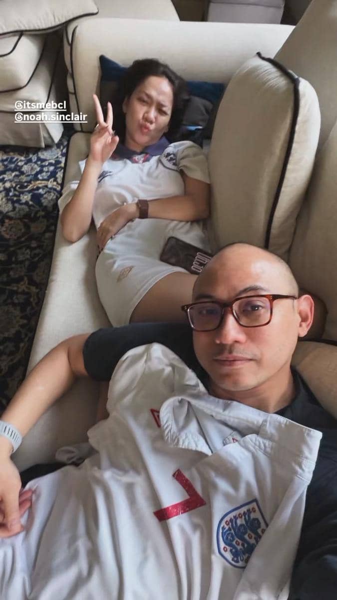 Dua orang mengenakan jersey tim nasional Inggris sedang bersantai di sofa, salah satunya berpose dengan tanda damai.