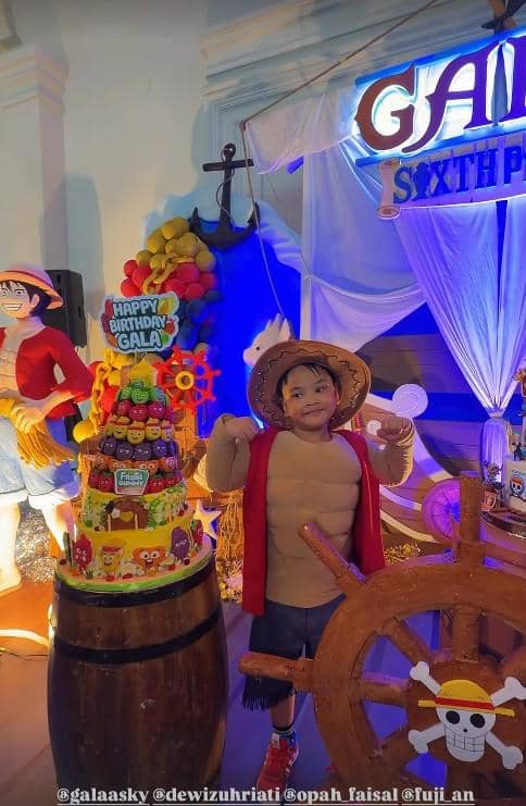 Seorang anak mengenakan kostum karakter bajak laut berdiri di samping kue ulang tahun bertema One Piece dengan dekorasi warna-warni.