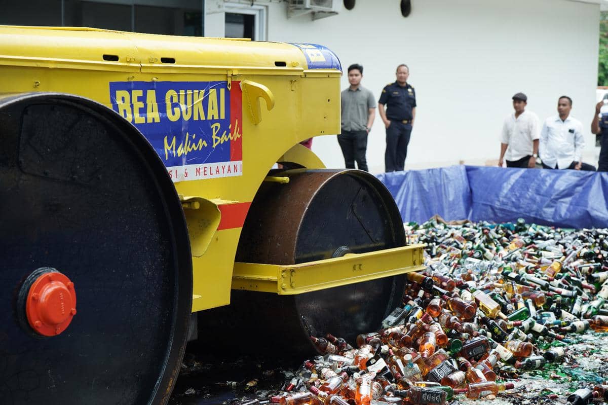 Alat berat Bea Cukai menghancurkan botol minuman keras hasil sitaan di area terbuka dengan beberapa petugas mengawasi di belakang.