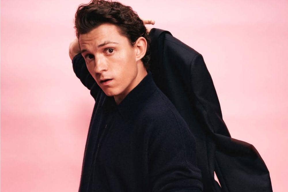 Tom Holland berpose dengan mengenakan kemeja gelap sambil memegang jaket di bahunya di depan latar belakang berwarna merah muda.