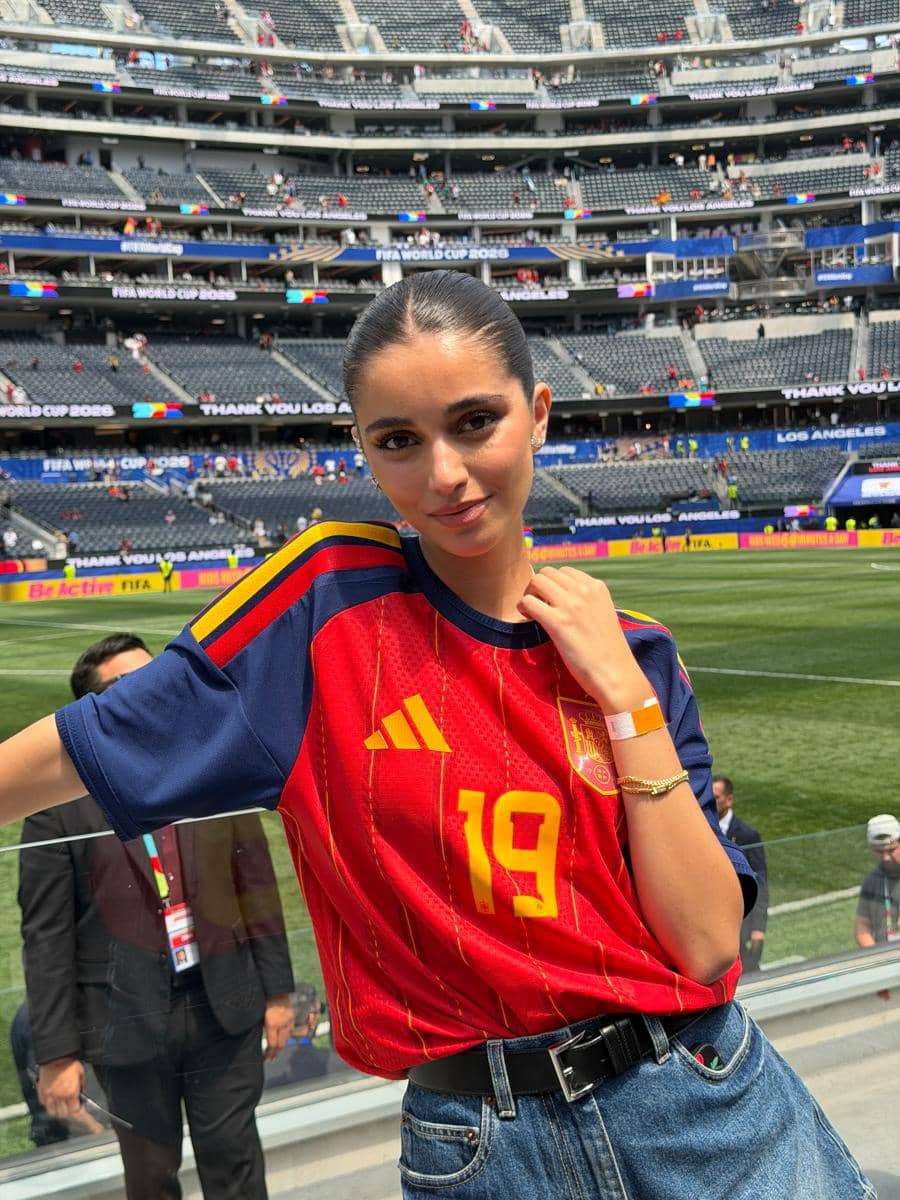 Seorang perempuan mengenakan jersey tim nasional Spanyol bernomor 19 berpose di stadion sepak bola dengan tribun penonton di latar belakang.