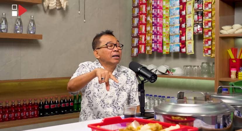 Seorang pria berkacamata mengenakan kemeja putih bermotif duduk di depan mikrofon di ruangan dengan latar rak berisi minuman dan kemasan produk.