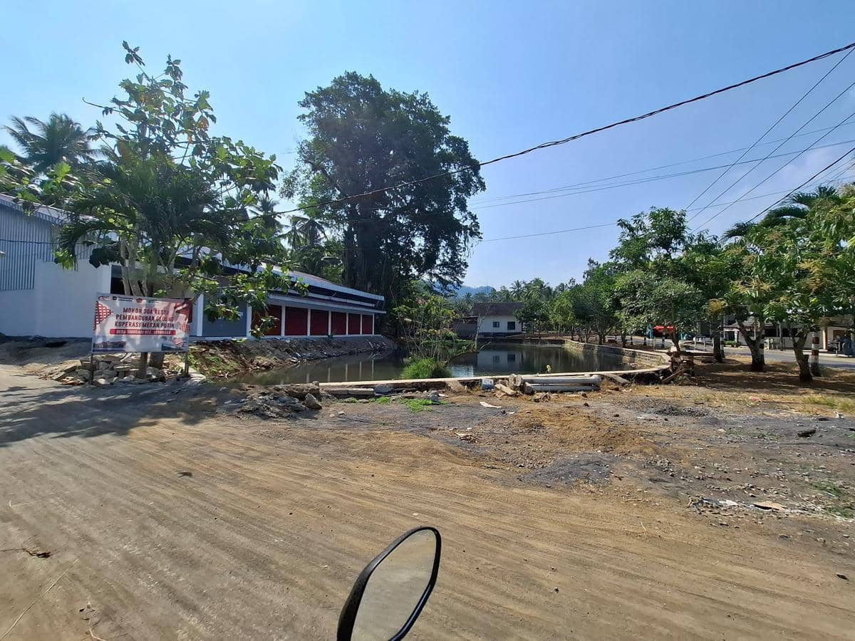 Bangunan KDKMP Desa Sidodadi berdiri di tepi kolam dengan pepohonan rindang dan jalan desa di bawah langit cerah.