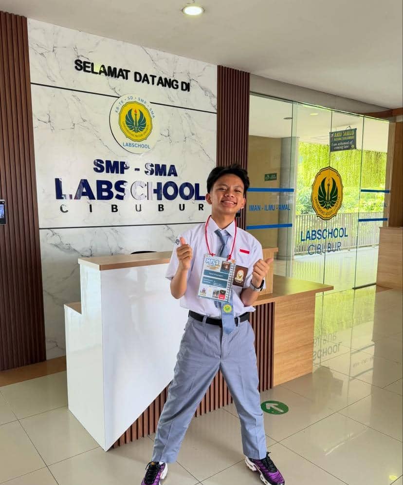 Seorang siswa berseragam putih abu-abu berpose ceria di lobi SMP-SMA Labschool Cibubur dengan tanda pengenal MPLS di lehernya.