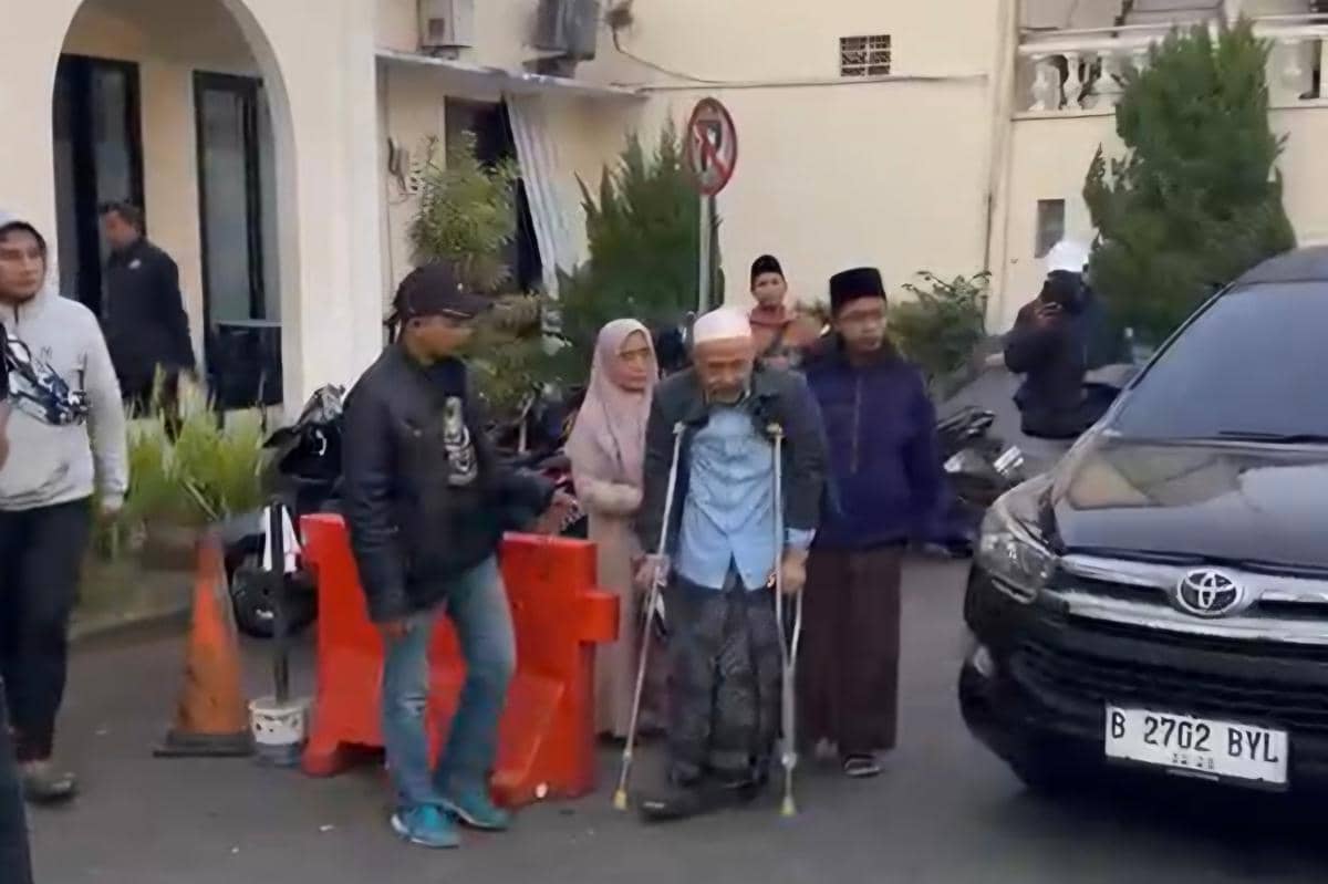 Seorang pria lanjut usia menggunakan kruk didampingi beberapa orang di depan sebuah bangunan dan mobil hitam di area parkir.