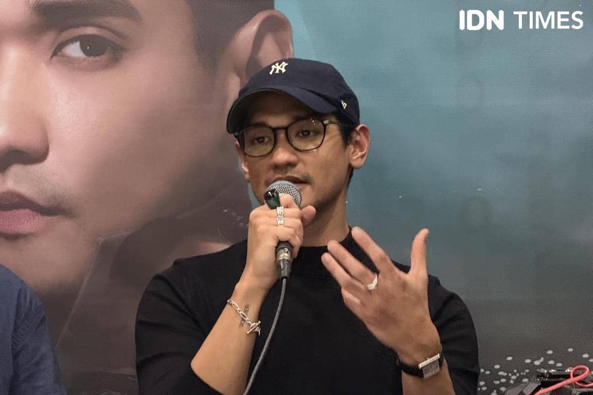 Seorang pria mengenakan topi dan kacamata berbicara sambil memegang mikrofon di depan poster besar wajahnya saat konferensi pers.