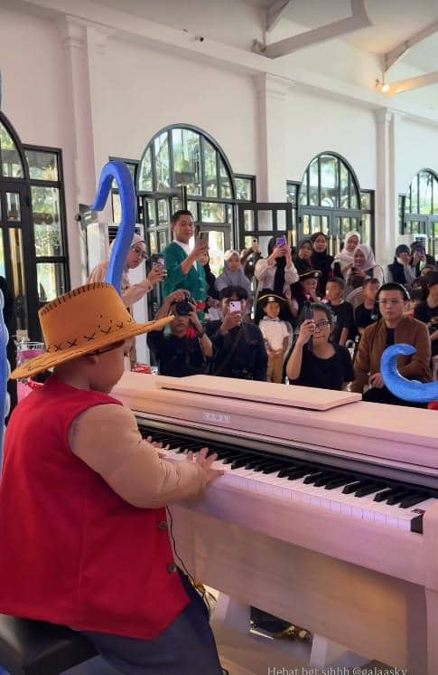 Seorang anak mengenakan kostum topi jerami dan rompi merah sedang bermain piano di depan tamu yang bertepuk tangan dan merekam momen tersebut.
