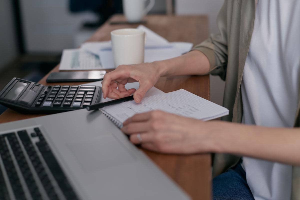 Seseorang menghitung angsuran kredit menggunakan kalkulator sambil mencatat di buku di meja kerja dengan laptop dan kopi.