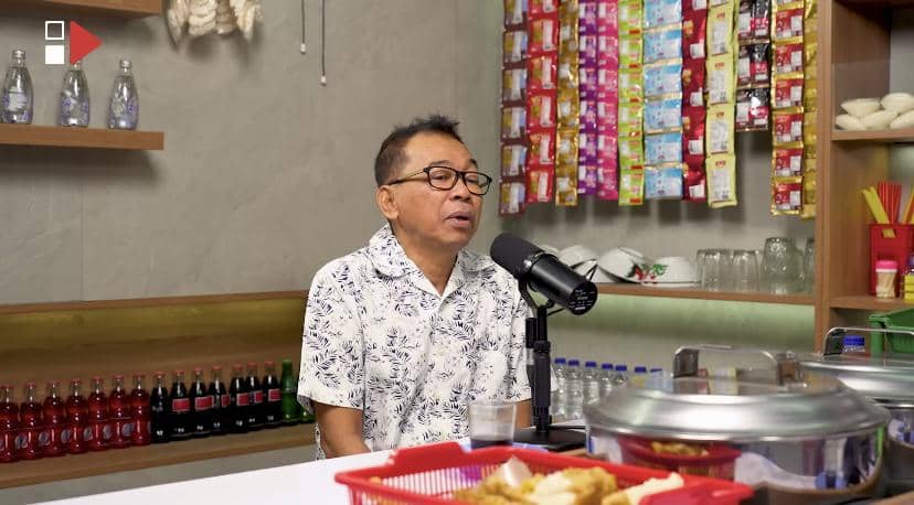 Seorang pria berkacamata mengenakan kemeja motif duduk di depan mikrofon di sebuah ruangan dengan latar rak minuman dan bumbu dapur.