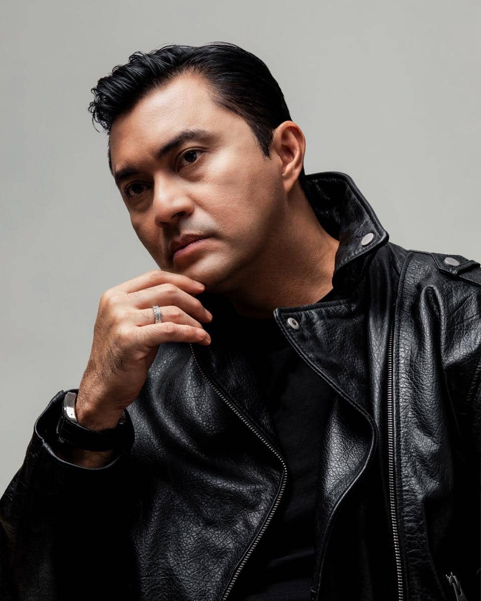 Seorang pria berpose dengan ekspresi serius mengenakan jaket kulit hitam dan kaus hitam di latar belakang abu-abu polos.