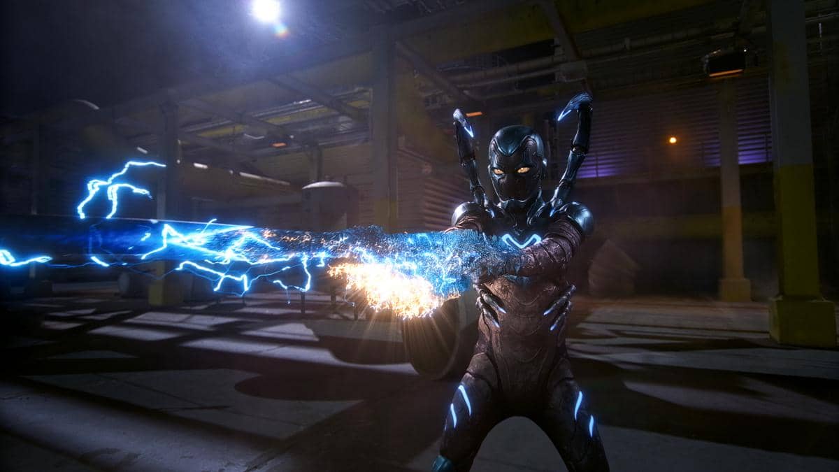Karakter superhero Blue Beetle mengenakan armor biru metalik memancarkan energi listrik biru di dalam area industri gelap.