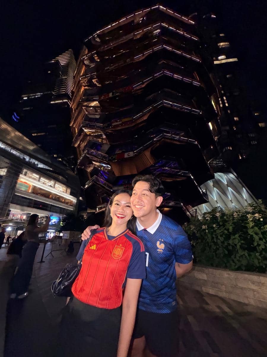 Seorang perempuan dan laki-laki berpose tersenyum di malam hari di depan bangunan The Vessel, New York, dengan pencahayaan kota yang terang.