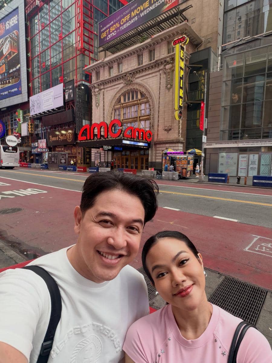 Seorang pria dan wanita berpose tersenyum di depan gedung bioskop AMC Empire di kawasan kota New York yang ramai dengan papan reklame.