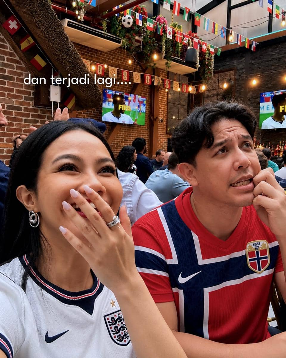Dua orang mengenakan jersey tim nasional Inggris dan Norwegia menonton pertandingan sepak bola di kafe dengan dekorasi bendera negara.