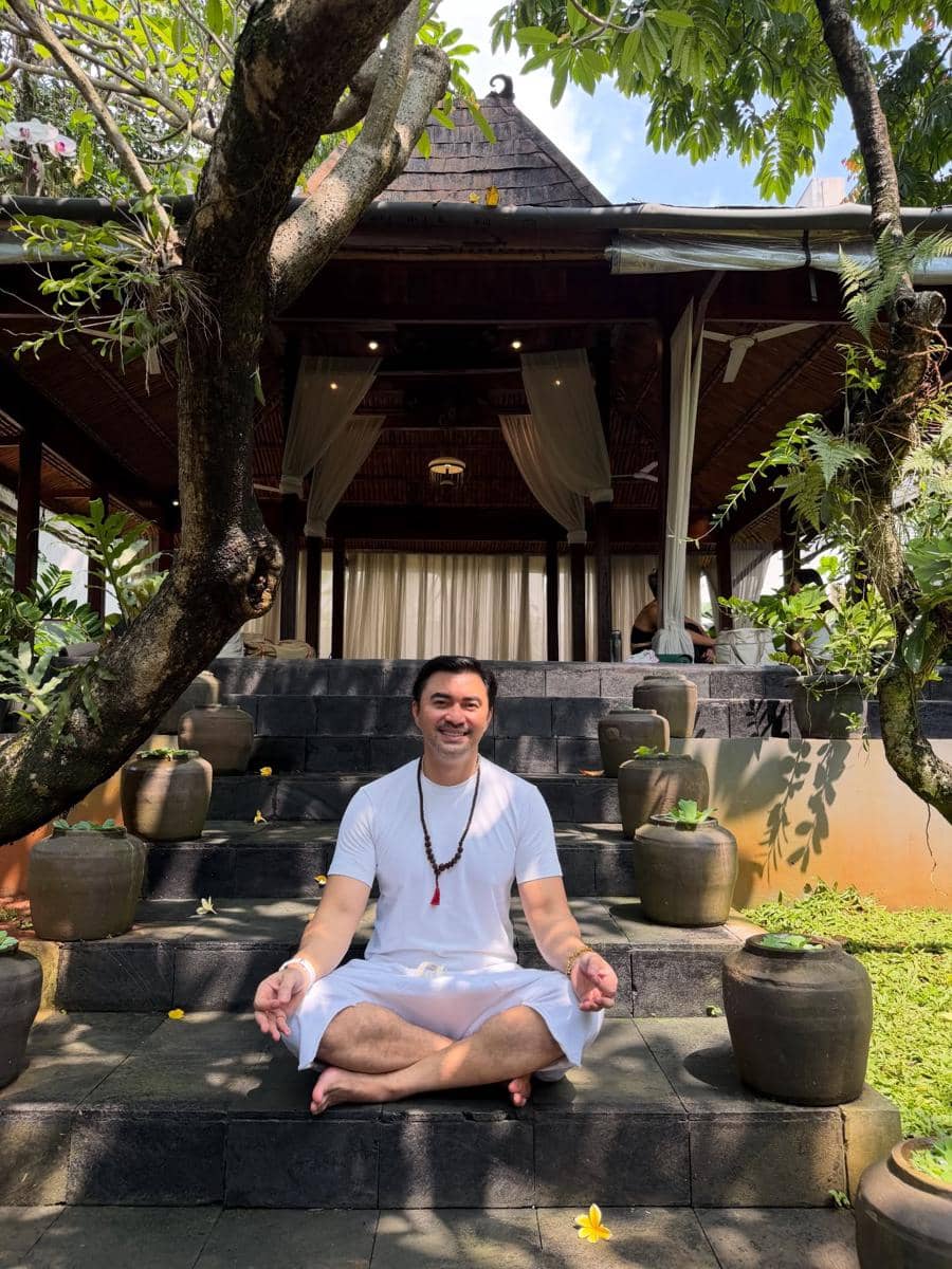 Seorang pria berpakaian putih duduk bersila sambil bermeditasi di tangga batu depan bangunan bergaya tradisional dengan taman hijau di sekitarnya.