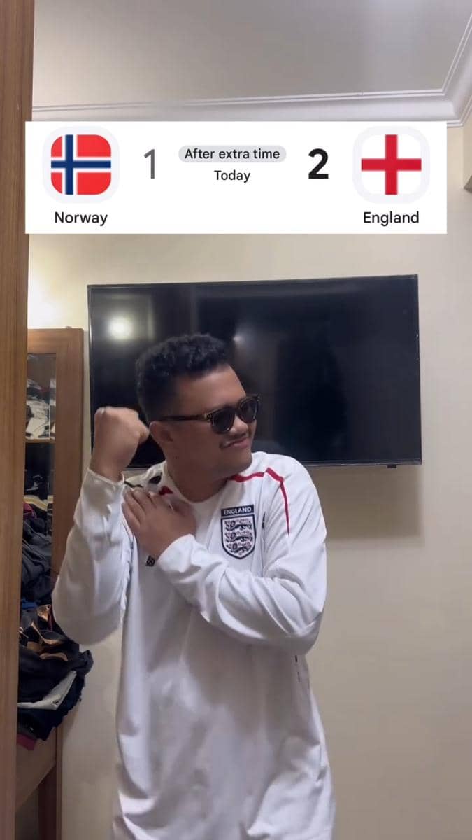 Seorang pria mengenakan jersey tim nasional Inggris berpose dengan gaya bangga di depan televisi yang menampilkan skor Inggris menang 2-1 atas Norwegia.