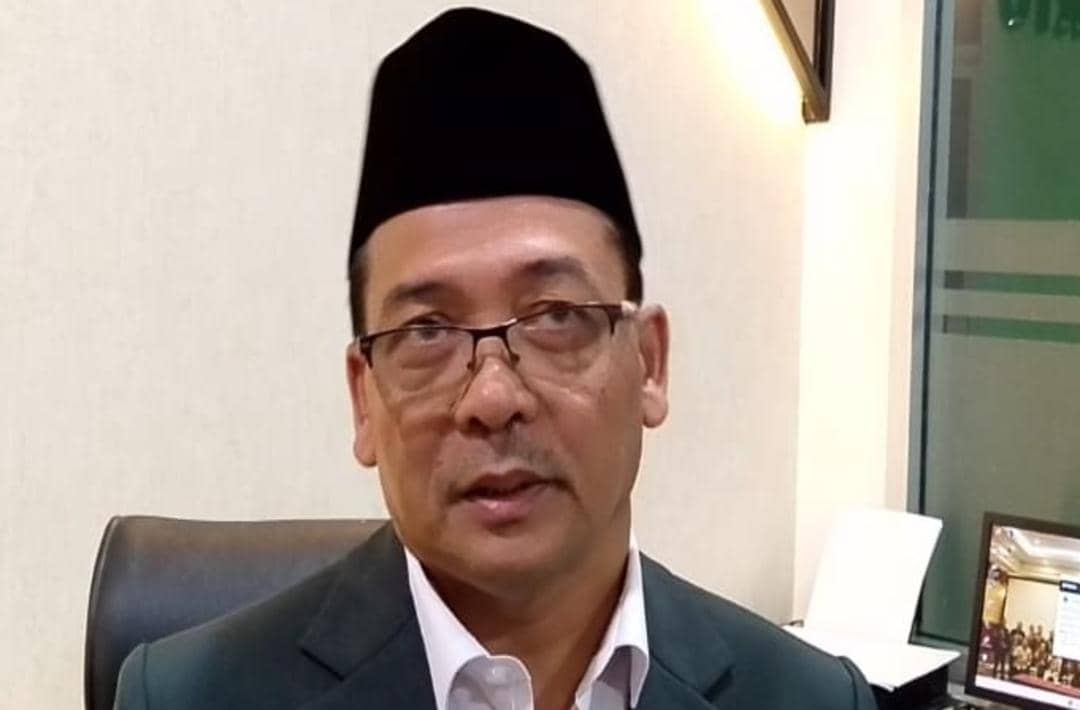 Seorang pria mengenakan jas gelap, kemeja putih, dan peci hitam sedang duduk di ruang kerja dengan latar dinding krem dan meja di belakangnya.