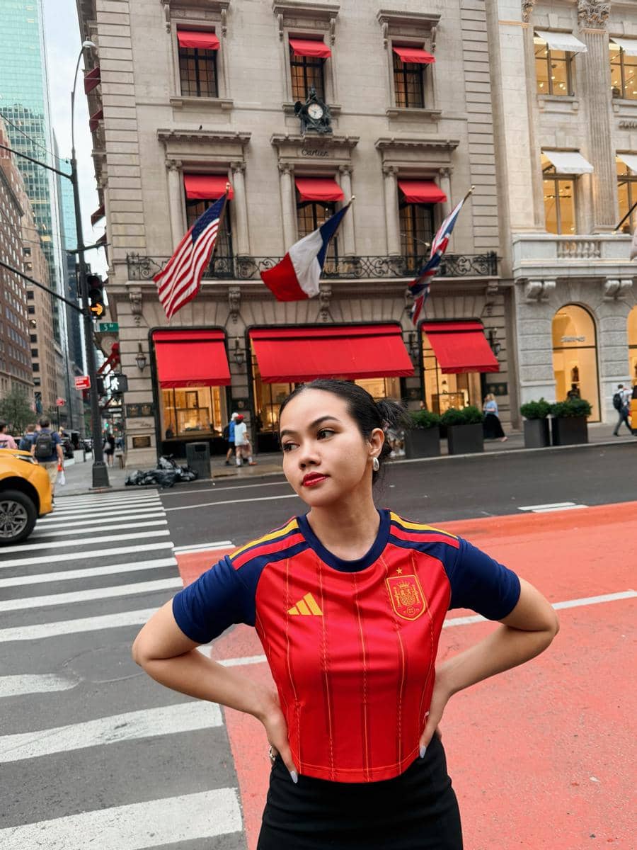 Seorang perempuan mengenakan jersey merah berdiri di persimpangan jalan kota New York dengan latar gedung bergaya klasik dan bendera Amerika.