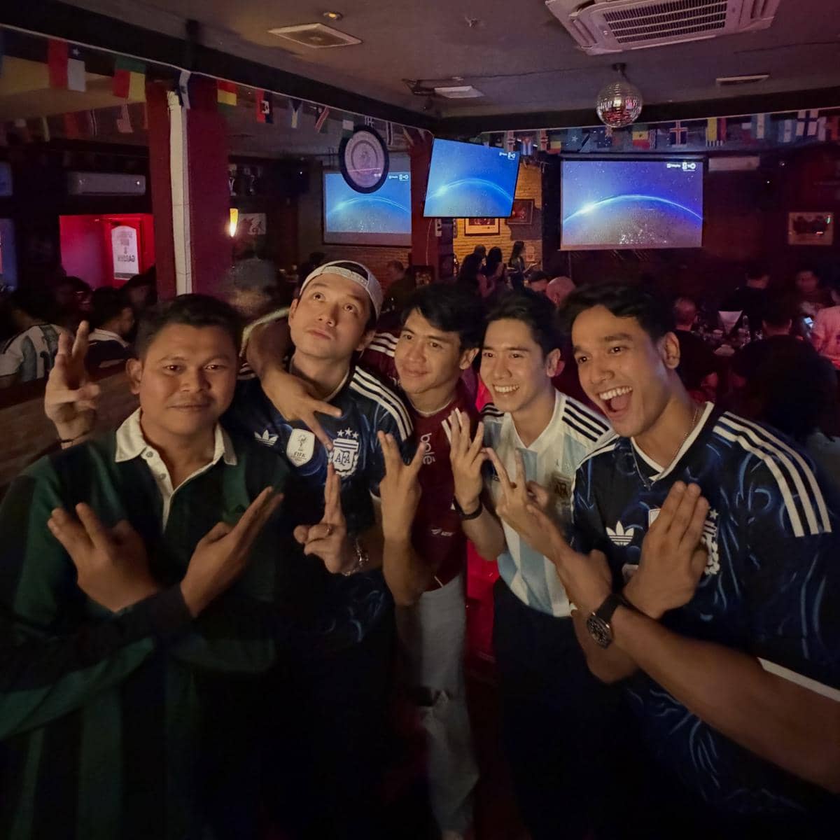Sekelompok pria mengenakan jersey sepak bola berpose bersama di sebuah bar saat menonton pertandingan Piala Dunia 2026.