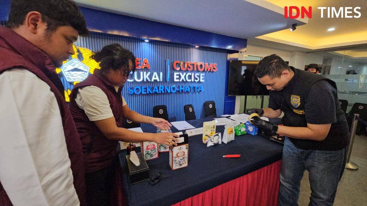 Petugas Bea Cukai Soekarno-Hatta memeriksa barang bukti bahan baku Happy Water di meja pemeriksaan dengan latar belakang logo Bea Cukai.