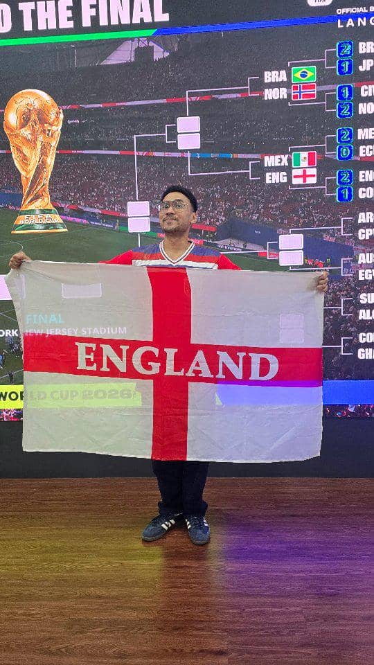 Seorang pria mengenakan jersey sepak bola memegang bendera Inggris di depan layar besar bergambar trofi Piala Dunia 2026.