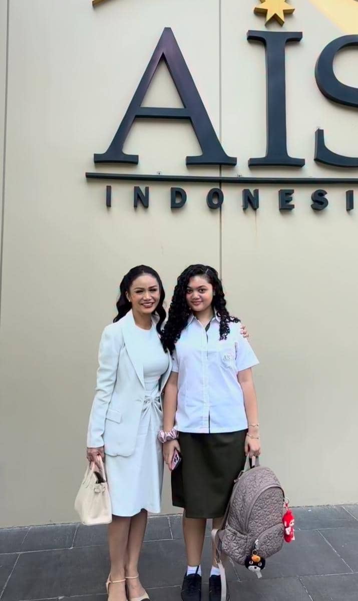 Seorang wanita dewasa berbusana putih berdiri di samping remaja berseragam sekolah dengan tas ransel di depan dinding bertuliskan AIS Indonesia.
