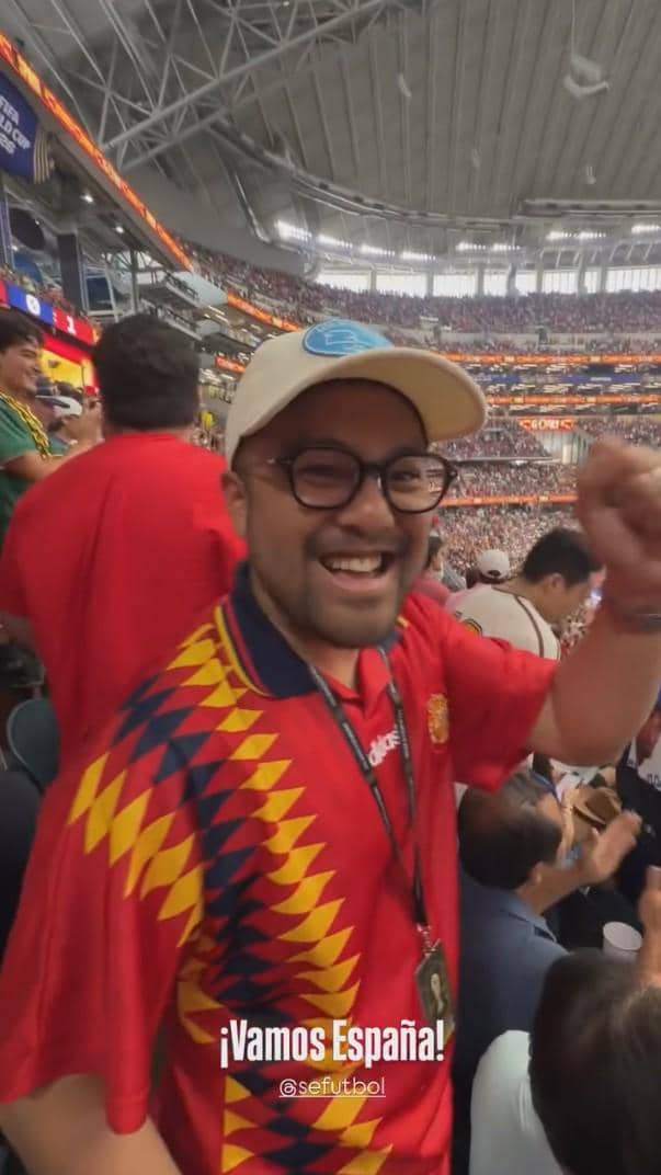 Seorang penonton mengenakan jersey tim Spanyol tersenyum dan mengangkat tangan di stadion penuh penonton saat pertandingan Spanyol versus Prancis.