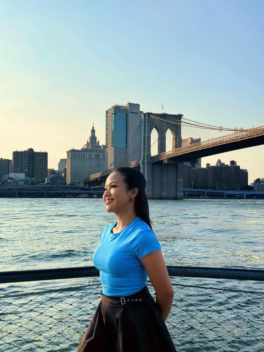 Seorang wanita mengenakan kaus biru dan rok hitam tersenyum di tepi sungai dengan latar Jembatan Brooklyn dan gedung-gedung New York.